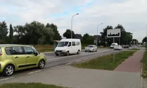 Zielony samochód i biały bus na drodze w terenie zabudowanym. W tle drzewa i domy.