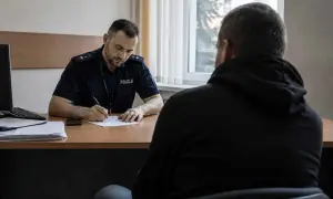 Policjant w mundurze pisze coś na papierze, siedząc naprzeciwko mężczyzny w bluzie z kapturem.