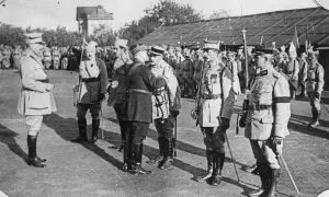 Oficerowie w mundurach, jeden z nich jako jeniec wojenny, stoją na placu przed budynkiem.