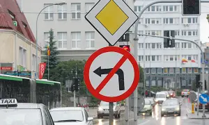 Znak zakazu skrętu w lewo na mokrej ulicy. Obok znak ustąp pierwszeństwa.