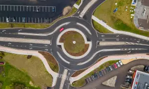 Widok z lotu ptaka na rondo z czerwonym samochodem. Widać parkingi, drogi z pasami ruchu i białe linie, a także zielone tereny. Kierunkowskaz jest widoczny na drodze prowadzącej do ronda.