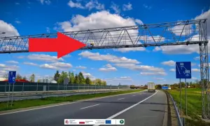 Na A4, pod niebieskim niebem z chmurami, widać bramownicę z urządzeniem do odcinkowego pomiaru prędkości. Czerwona strzałka wskazuje na system monitorowania.