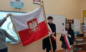 Uczniowie tworzą poczet sztandarowy, prezentując biało-czerwoną flagę Polski. Dziewczyna czyta tekst, chłopiec stoi obok.
