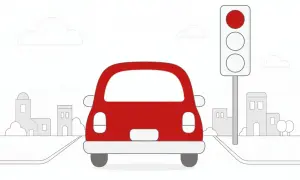 Czerwony samochód zatrzymał się na skrzyżowaniu przed czerwonym światłem. W tle widać budynki i chmury.