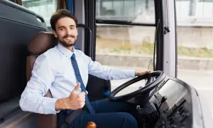 Uśmiechnięty kierowca autobusu, w białej koszuli i granatowym krawacie, pokazuje kciuk w górę. Jego czas pracy przebiega spokojnie.