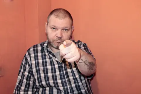 Mężczyzna w kratę wskazuje palcem, na ramieniu tatuaż z Indianinem. "234 kk" - czy to jego numer?