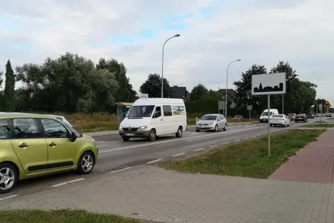 Zielony samochód i biały bus na drodze w terenie zabudowanym. W tle drzewa i domy.