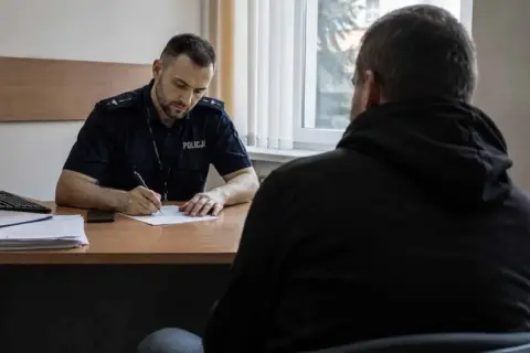 Policjant w mundurze pisze coś na papierze, siedząc naprzeciwko mężczyzny w bluzie z kapturem.