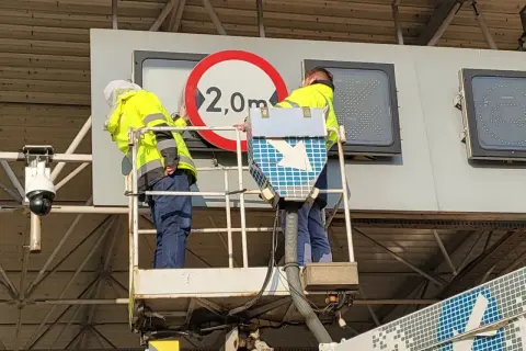 Pracownicy na podnośniku montują znak ograniczenia wysokości 2,0m przy bramkach viatoll.