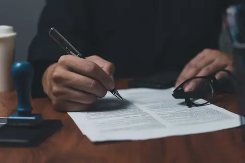 Dłoń podpisuje dokument piórem, obok okulary i pieczęć. To ważny list żelazny, potwierdzający umowę.