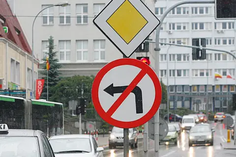 Znak zakazu skrętu w lewo na mokrej ulicy. Obok znak ustąp pierwszeństwa.