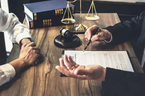Prawnik wskazuje piórem na umowę, podczas gdy klient trzyma ręce złożone na drewnianym stole. W tle widać młotek sędziowski i wagę sprawiedliwości. To zstępny etap negocjacji.