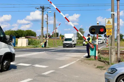 Pracownik obsługuje przejazd kolejowy z zaporami. Czerwone światło sygnalizatora i opuszczone bariery zatrzymują ruch pojazdów.