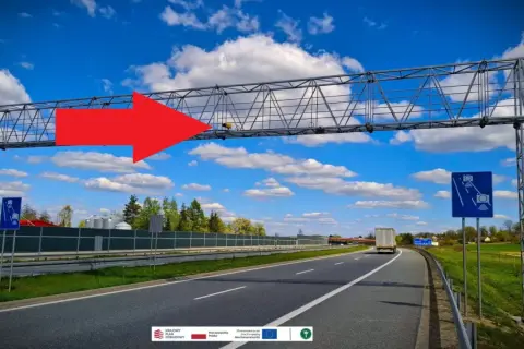 Na A4, pod niebieskim niebem z chmurami, widać bramownicę z urządzeniem do odcinkowego pomiaru prędkości. Czerwona strzałka wskazuje na system monitorowania.