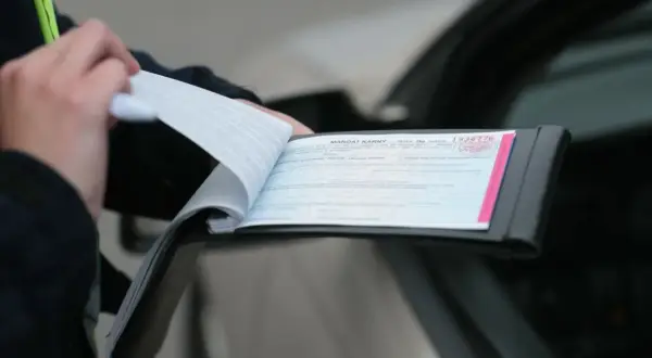 Policjant wypisuje mandat karny w notesie przy samochodzie.