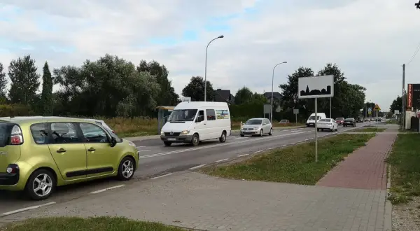 Zielony samochód i biały bus na drodze w terenie zabudowanym. W tle drzewa i domy.