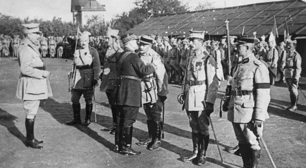 Oficerowie w mundurach, jeden z nich jako jeniec wojenny, stoją na placu przed budynkiem.
