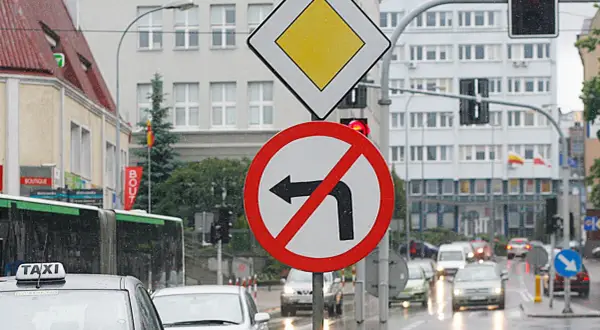 Znak zakazu skrętu w lewo na mokrej ulicy. Obok znak ustąp pierwszeństwa.