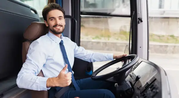 Uśmiechnięty kierowca autobusu, w białej koszuli i granatowym krawacie, pokazuje kciuk w górę. Jego czas pracy przebiega spokojnie.