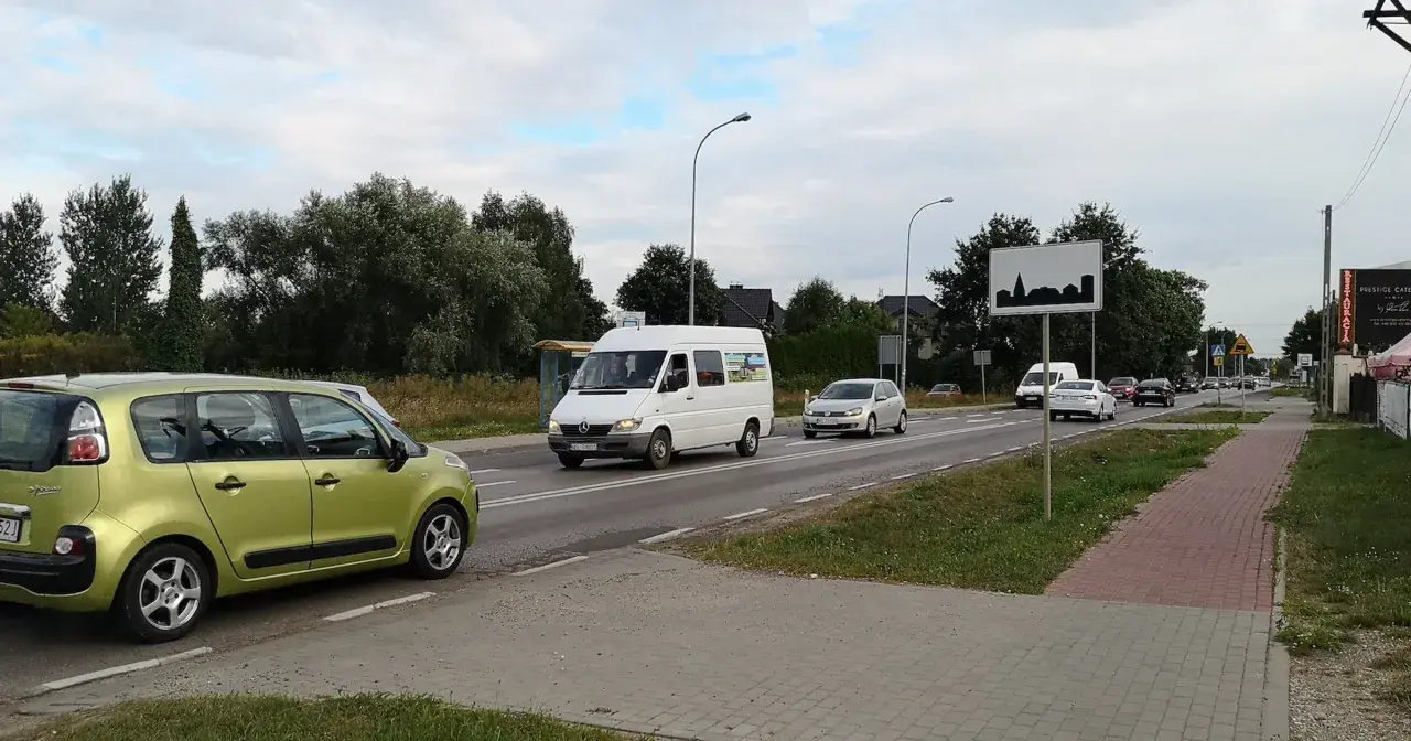 Zielony samochód i biały bus na drodze w terenie zabudowanym. W tle drzewa i domy.