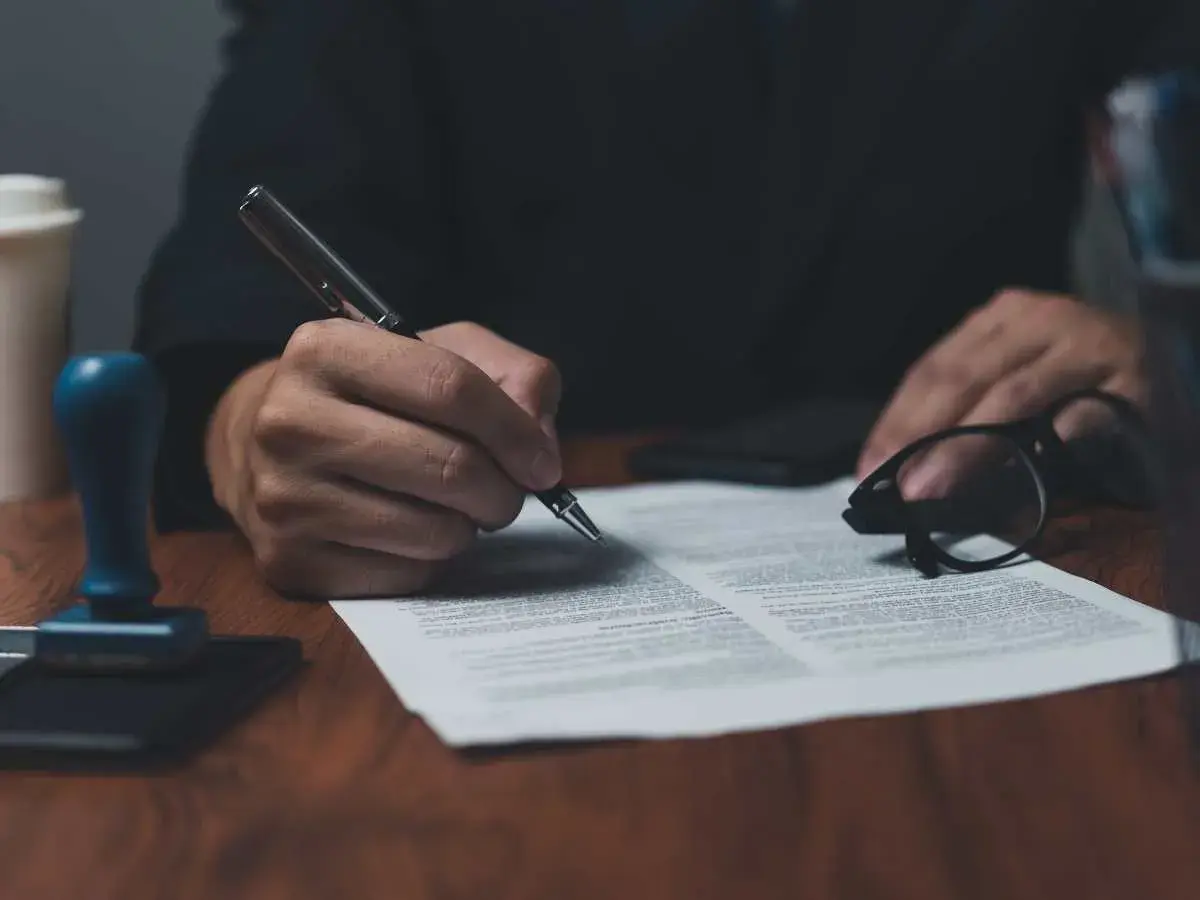Dłoń podpisuje dokument piórem, obok okulary i pieczęć. To ważny list żelazny, potwierdzający umowę.