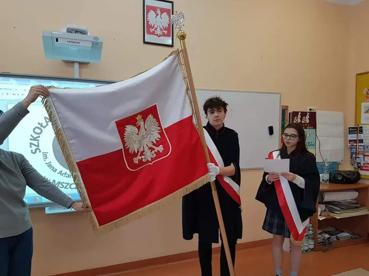 Uczniowie tworzą poczet sztandarowy, prezentując biało-czerwoną flagę Polski. Dziewczyna czyta tekst, chłopiec stoi obok.