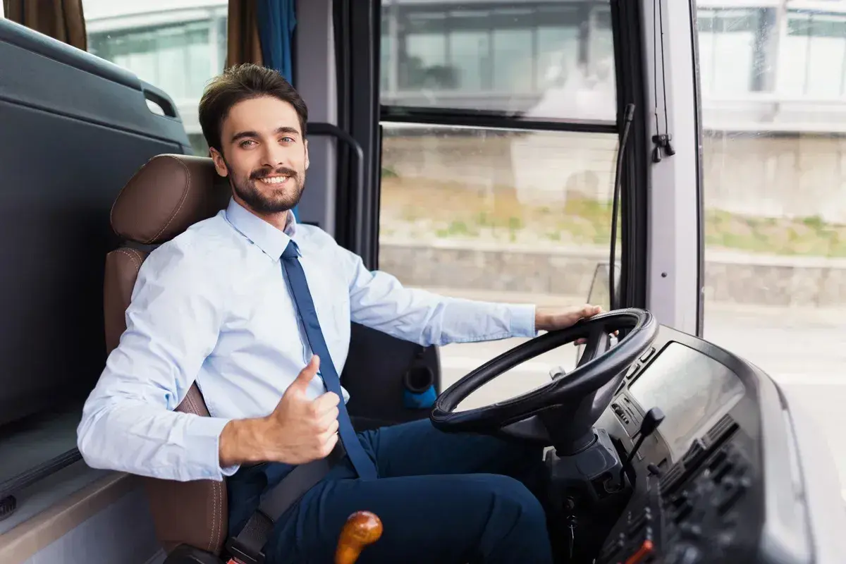Uśmiechnięty kierowca autobusu, w białej koszuli i granatowym krawacie, pokazuje kciuk w górę. Jego czas pracy przebiega spokojnie.