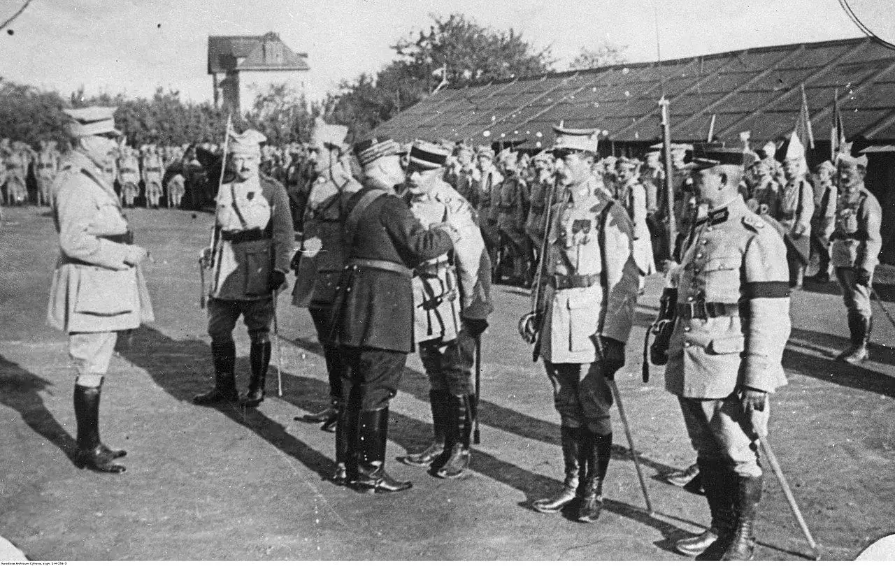 Oficerowie w mundurach, jeden z nich jako jeniec wojenny, stoją na placu przed budynkiem.