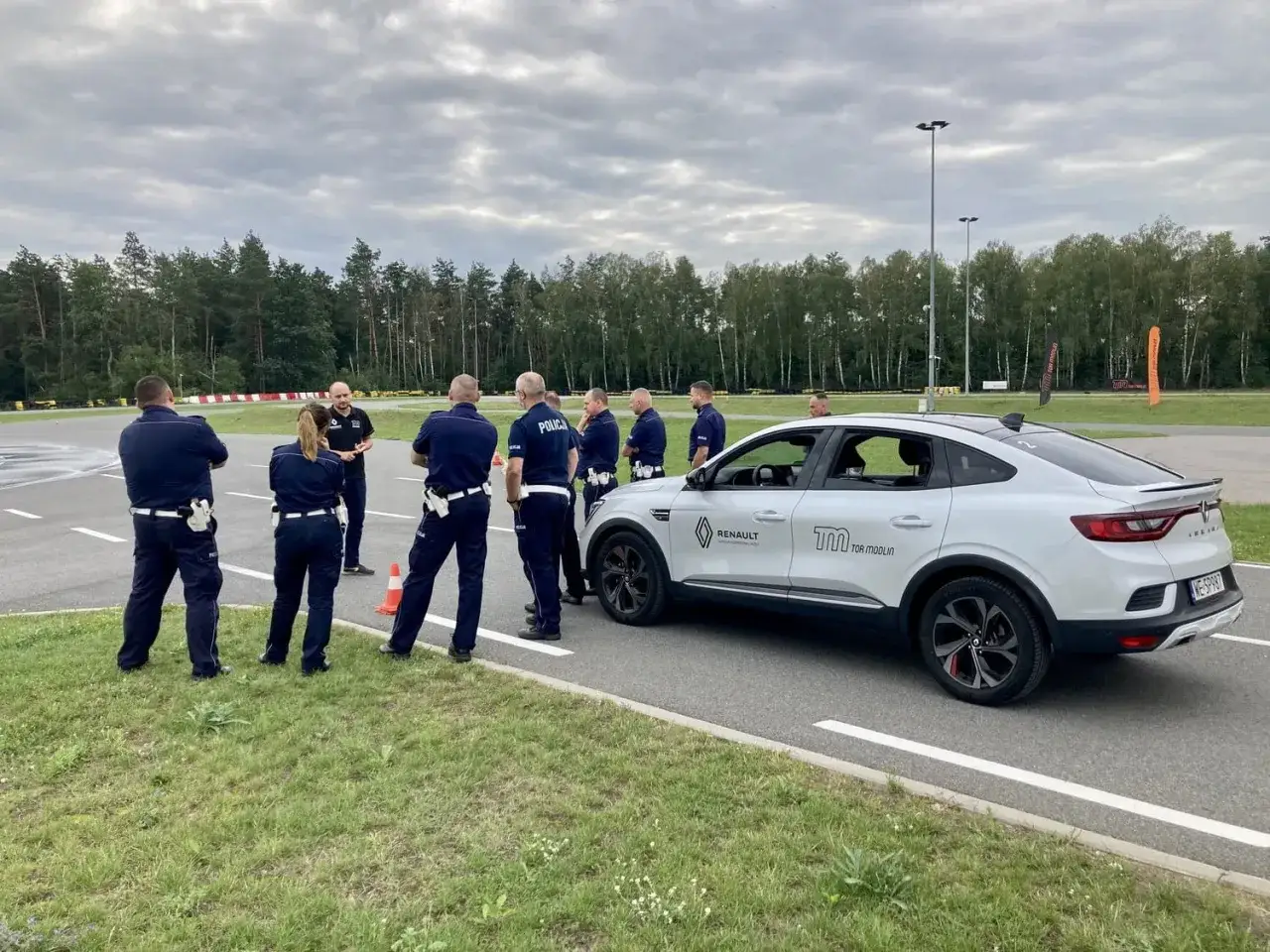 Policjanci na torze szkoleniowym podczas kursu na pojazdy uprzywilejowane. Instruktor omawia zadania przy białym Renault.