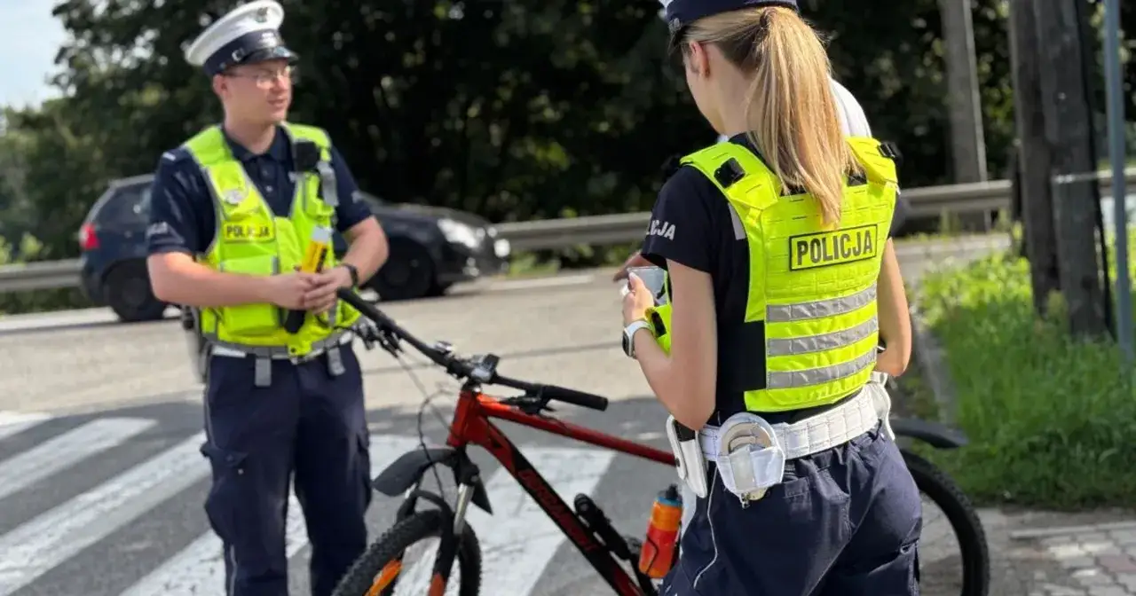 Policjanci kontrolują rowerzystę, który mógł mieć jazda rowerem po alkoholu. Na drodze stoi rower, a funkcjonariusze mają na sobie kamizelki odblaskowe.