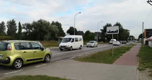 Zielony samochód i biały bus na drodze w terenie zabudowanym. W tle drzewa i domy.