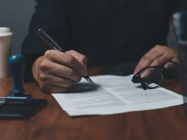 Dłoń podpisuje dokument piórem, obok okulary i pieczęć. To ważny list żelazny, potwierdzający umowę.