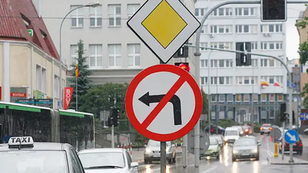 Znak zakazu skrętu w lewo na mokrej ulicy. Obok znak ustąp pierwszeństwa.