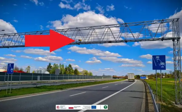 Na A4, pod niebieskim niebem z chmurami, widać bramownicę z urządzeniem do odcinkowego pomiaru prędkości. Czerwona strzałka wskazuje na system monitorowania.